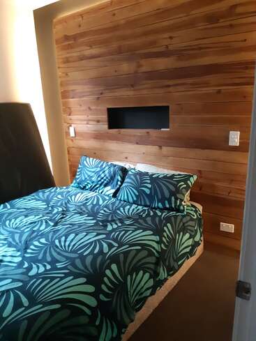 A imagem mostra um quarto com uma parede de madeira que apresenta uma área rebaixada, uma cama adornada com um edredom com estampa de folhas azuis e verdes e uma cabeceira preta.