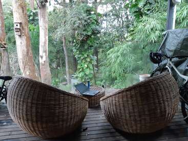 A imagem retrata um ambiente externo sereno com duas cadeiras de vime em um deck de madeira, cercado por vegetação exuberante e árvores, exalando uma sensação de tranquilidade e relaxamento.