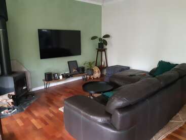 A imagem mostra uma sala de estar aconchegante com um sofá de couro escuro, piso de madeira e paredes verdes, com uma TV na parede e uma lareira.