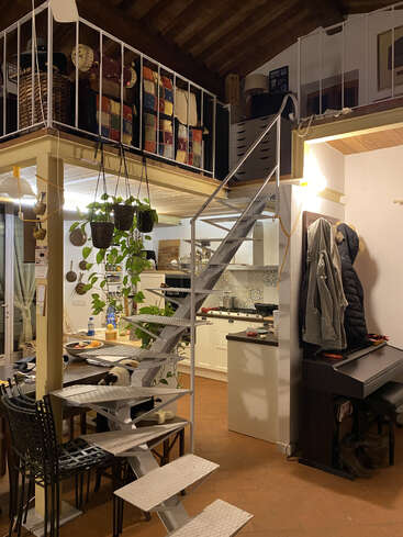 Um loft aconchegante e eclético com uma escada de metal, plantas penduradas, almofadas coloridas de patchwork, cozinha, mesa de jantar, piano, casacos pendurados e um gato relaxando embaixo.