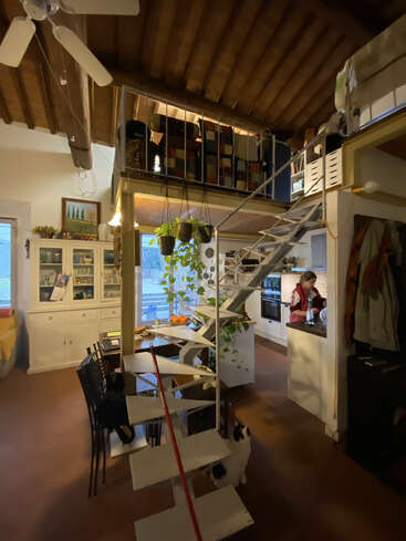 Um loft aconchegante e rústico com teto de madeira, móveis brancos, plantas e uma escada em espiral. Uma mulher cozinha na cozinha enquanto um gato fica sentado embaixo.