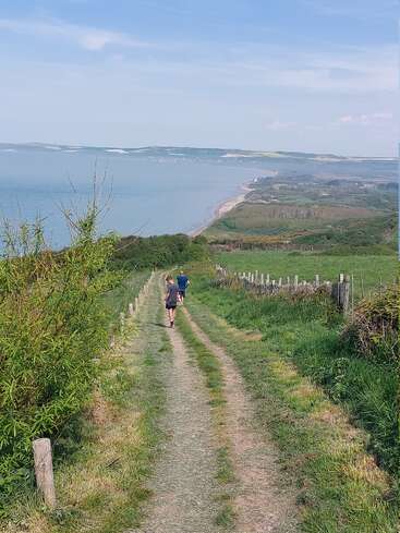 Two children walk along a grassy coastal path bordered by fences, overlooking the sea and cliffs under a clear sky, surrounded by lush greenery and nature.
