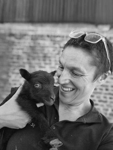 A smiling person wearing glasses on their head lovingly holds a small, dark-colored baby goat. The black and white image captures a joyful, tender moment.