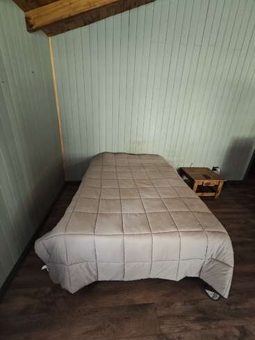 Une petite chambre confortable comprend un lit simple avec un édredon beige, un sol en bois, des panneaux muraux verdâtres, un plafond en bois et une petite table d'appoint. Arrangement simple.