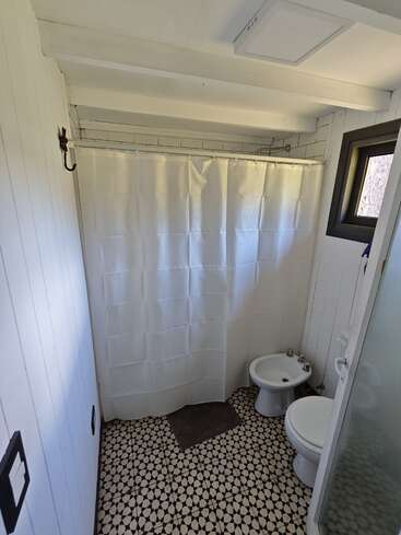 Cette salle de bains comprend un sol carrelé à motifs, des murs blancs, une douche avec rideau, des toilettes, un bidet, une petite fenêtre et un tapis de bain marron.