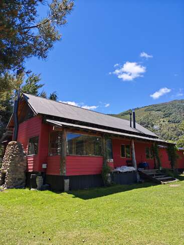 Une cabane en bois rouge avec une cheminée en pierre est posée sur de l'herbe verte, entourée d'arbres et de collines, sous un ciel bleu vif avec des nuages épars.