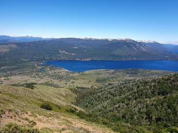 Un paysage magnifique représentant un lac bleu clair, entouré de forêts vertes, de collines ondulantes et de montagnes lointaines sous un ciel bleu lumineux et sans nuages. Paisible et serein.
