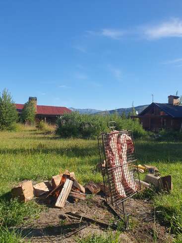 Dans un champ herbeux, de la viande est cuite sur un gril au-dessus d'un feu ouvert. Deux maisons rustiques et des montagnes lointaines sont visibles sous un ciel dégagé.