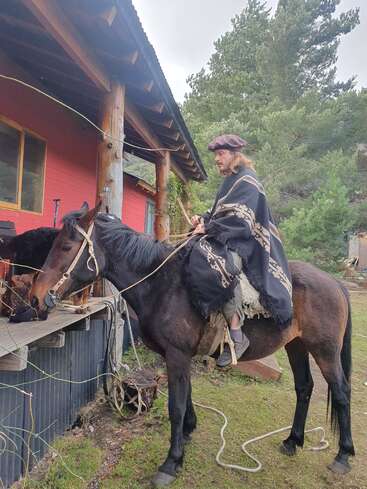 Un homme portant des vêtements traditionnels et un béret monte un cheval noir près d'une maison rustique en bois, entourée d'arbres verts et d'un paysage naturel en plein air.