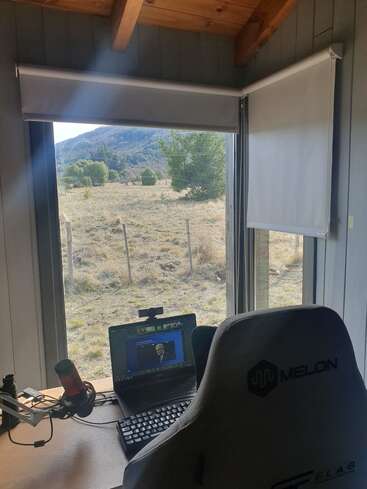 Un espace de travail confortable avec une chaise de jeu, un microphone, un ordinateur portable et un clavier fait face à de grandes fenêtres offrant une vue pittoresque sur la campagne avec des collines, des arbres et de la lumière naturelle.