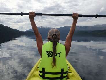 Une personne est assise dans un kayak jaune, tenant une pagaie triomphante au-dessus de sa tête, portant un gilet de sécurité brillant, entourée d'un lac aux eaux calmes et de montagnes. Une scène magnifique et paisible.