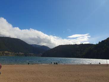 Scène de lac paisible avec plage de sable, collines vertes et montagnes lointaines. Des personnes se détendent sur le rivage sous un ciel bleu clair et lumineux avec des nuages épars.