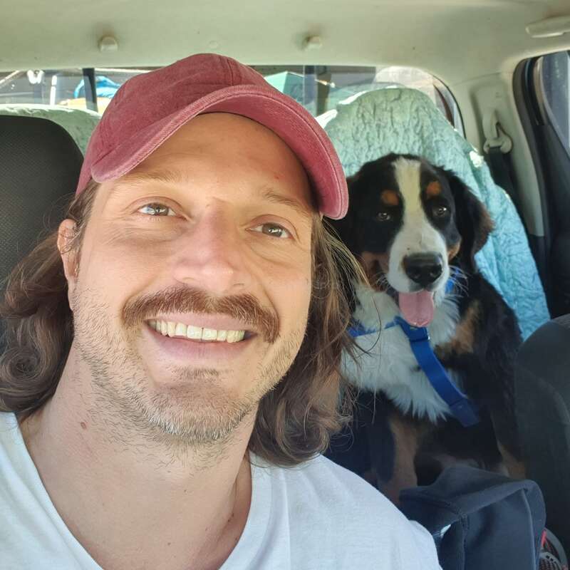 A man with a mustache and red cap smiles for a selfie inside a car while his happy Bernese Mountain Dog sits in the backseat, tongue out.