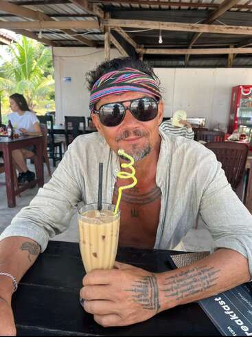 A man with tattoos, sunglasses, and a colorful headband sits at a café table, holding an iced coffee with a curly straw, enjoying a relaxed atmosphere.