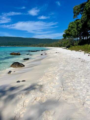 A pristine beach with soft white sand, turquoise waters, scattered rocks, lush green trees, distant hills, and a vibrant blue sky with wispy clouds overhead.