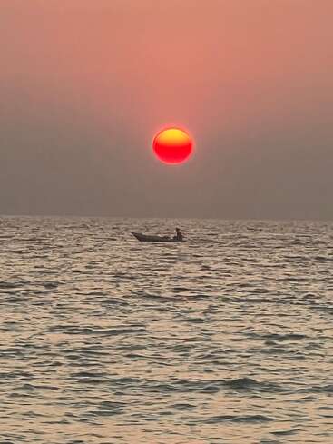 A lone person in a small boat floats on calm ocean waters, beneath a striking, vibrant red and yellow sunset, creating a serene, peaceful scene.
