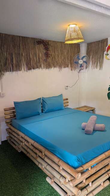 This cozy room features a bamboo bed with blue sheets, faux grass flooring, a wicker lamp, fan, thatched wall decor, and neatly rolled towels. Tropical vibe.