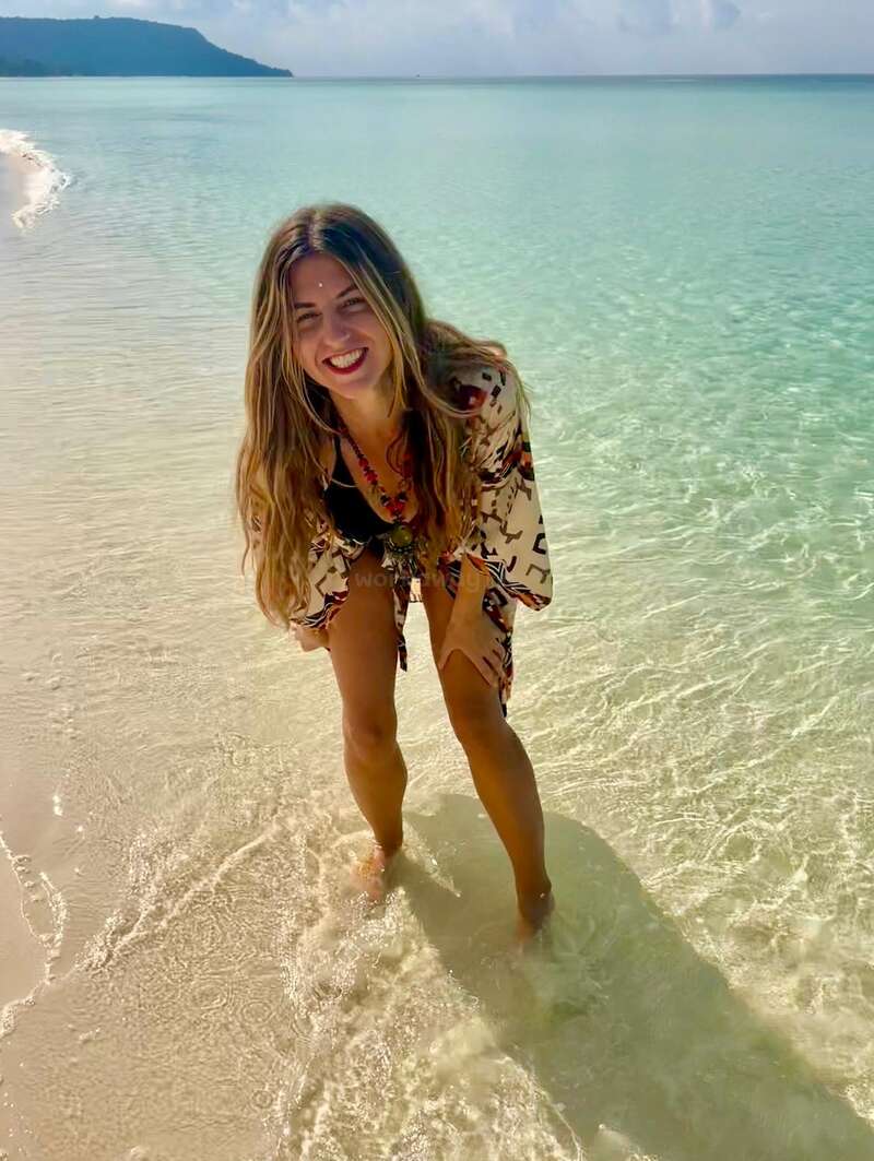 A smiling woman stands in shallow, clear turquoise water on a sunny beach, wearing a patterned cover-up. The scenery is tranquil, with gentle waves and distant hills.