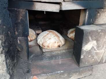 Três pães redondos estão assando dentro de um forno tradicional de pedra ou tijolo. O pão está douradinho, e o forno parece rústico e bastante usado.