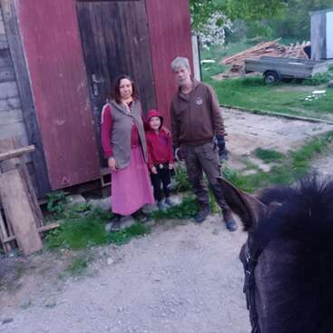 Uma família de três pessoas está do lado de fora de um galpão rústico de madeira, posando ao lado de um cavalo, com grama verde e pedaços de madeira espalhados no fundo rural.