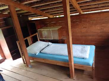 Esta imagem mostra um quarto simples de madeira com teto de vigas à vista, uma cama de solteiro com roupa de cama azul, prateleiras e um pequeno armário na parede.