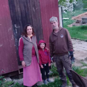 Uma família de três pessoas está do lado de fora de um galpão rústico de madeira, posando ao lado de um cavalo, com grama verde e pedaços de madeira espalhados no fundo rural.