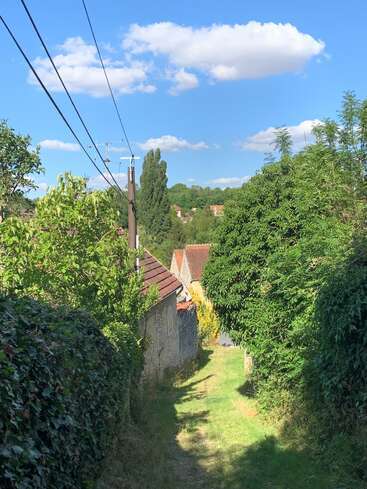 Un estrecho camino de hierba serpentea entre frondosos setos y casitas de piedra, bajo cables de alta tensión y un cielo azul brillante salpicado de esponjosas nubes blancas. Una apacible escena rural.