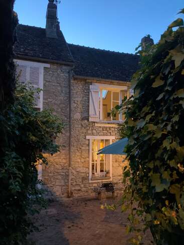 Una encantadora casa de piedra con contraventanas blancas está cálidamente iluminada desde el interior. La hiedra enmarca el patio, mientras el crepúsculo se instala, creando una acogedora y acogedora escena nocturna.