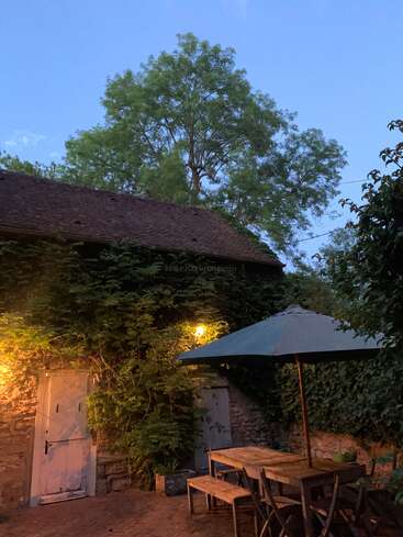 Un acogedor patio al aire libre con mesa y sillas de madera, una gran sombrilla, un muro de piedra cubierto de hiedra, luces brillantes, exuberante vegetación y un árbol alto bajo el cielo nocturno.