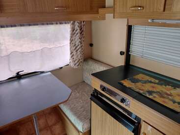 Esta imagem mostra o interior aconchegante de uma van de camping com uma mesinha, um banco almofadado, armários de madeira, fogão, geladeira e um tapete com estampa floral.
