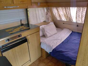 Esta imagem mostra o interior aconchegante de uma van de camping, com uma pequena cozinha compacta e uma cama bem arrumada com roupa de cama azul e branca ao lado das janelas.