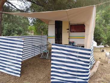 Uma pequena caravana com toldo está estacionada numa área arborizada, cercada por biombos listrados de azul e branco, cadeiras de jardim, uma mesa e um cachorro por perto.