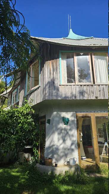 Uma casa charmosa de dois andares com revestimento de madeira, janelas amplas, detalhes em turquesa e um telhado curvo único. Cercada por uma vegetação exuberante, plantas e ferramentas de jardim do lado de fora.