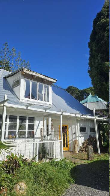 Uma charmosa casa branca com telhado de metal inclinado, porta amarela e janelas amplas, cercada por plantas verdes, sob um céu azul vibrante em um dia ensolarado.
