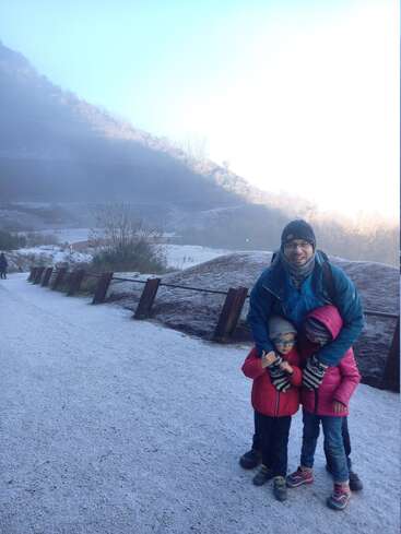 A man and two children in colorful winter clothing stand on a snowy path, smiling together. Misty, sunlit mountains and frosty scenery surround them.