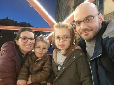 A family of four, all wearing glasses, smiles for a photo together. They are outdoors at dusk, with festive lights and buildings in the background.