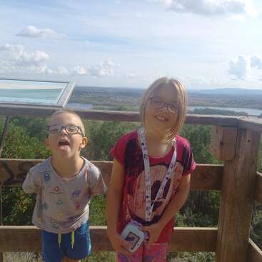 Two children with glasses stand on a wooden lookout platform. The boy is making a funny face while the girl smiles, with beautiful scenery behind them.