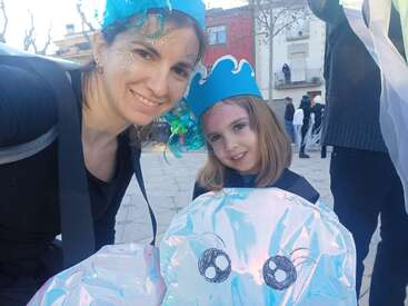 A woman and a girl are smiling at an outdoor event, wearing blue sea-themed costumes and crowns. The girl holds a shiny jellyfish craft.