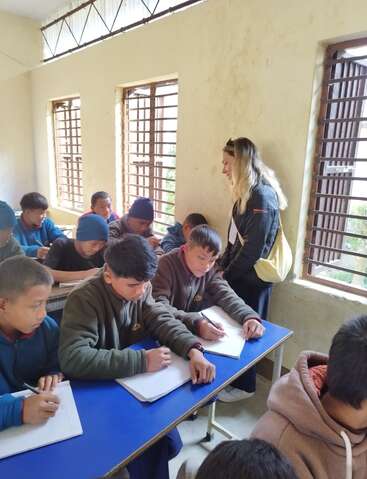 A classroom scene with several students sitting at blue desks, writing in notebooks, while a teacher stands nearby, observing and assisting. Sunlight streams through barred windows.