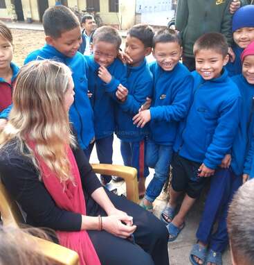 A group of smiling children in blue uniforms gather around a seated woman with blonde hair and a red scarf, laughing happily together outdoors.