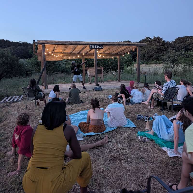 Um grupo de pessoas senta-se ao ar livre na grama e em cadeiras, assistindo a um artista em um palco de madeira sob luzes de corda durante um evento noturno na natureza.
