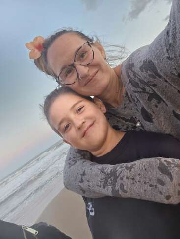Une femme et un enfant sourient joyeusement sur la plage. La femme porte des lunettes et une fleur dans les cheveux ; elle serre l'enfant contre elle pour le protéger des vagues.