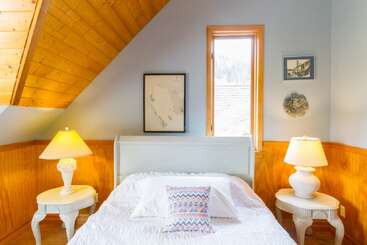 Dieses gemütliche Schlafzimmer hat ein weißes Bett mit gemusterten Kissen, zwei passende Lampen auf Beistelltischen, Holzakzente, eine schräge Decke und gerahmte Kunst an pastellfarbenen Wänden.