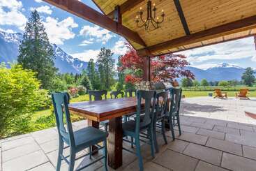 Auf der überdachten Terrasse steht ein hölzerner Esstisch mit blaugrünen Stühlen. Von hier aus hast du einen Blick auf saftig grünes Gras, leuchtende Bäume, Berggipfel und Adirondack-Stühle unter einem blauen Himmel.