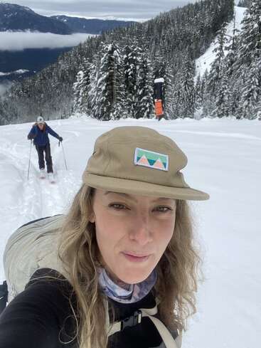 Eine Frau macht ein Selfie beim Wandern durch eine verschneite Berglandschaft. Eine andere Person folgt ihr; Tannenbäume und ferne Berge vervollständigen die schöne Winterszene.