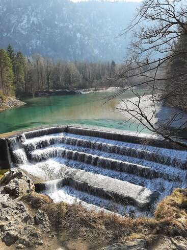 Das Bild zeigt einen ruhigen Wasserfall, der in einen stillen See stürzt, umgeben von üppigen Bäumen und einer majestätischen Bergkulisse in der Ferne.
