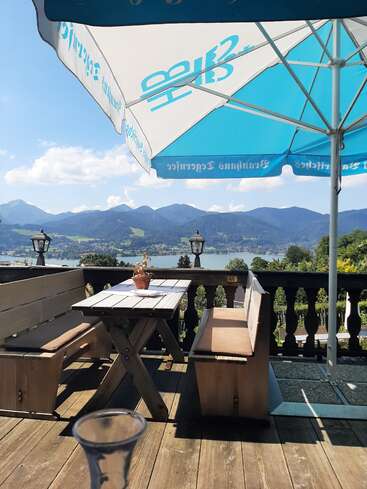 Das Bild zeigt eine ruhige Umgebung im Freien mit einer Holzterrasse, einem Tisch und einer Bank, einem blau-weißen Sonnenschirm und einer malerischen Aussicht auf Berge und einen See im Hintergrund.