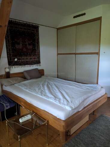 Esse quarto aconchegante tem uma cama de madeira com roupa de cama branca, dois travesseiros, tapeçaria de parede, abajur, pequena mesa de vidro, armário embutido e piso de madeira.
