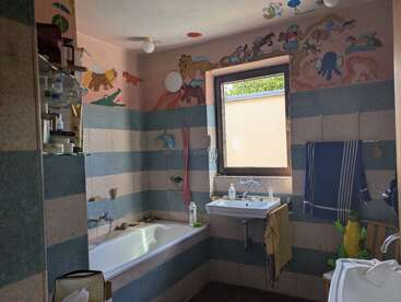 Este é um banheiro infantil com murais de parede coloridos com tema de animais, banheira, pia, janela, azulejos listrados, toalhas, prateleiras com produtos de higiene pessoal e luz natural.