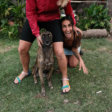 A imagem mostra duas mulheres e um cachorro em um cenário de quintal, com as mulheres ajoelhadas e em pé na grama, cercadas por plantas e árvores.